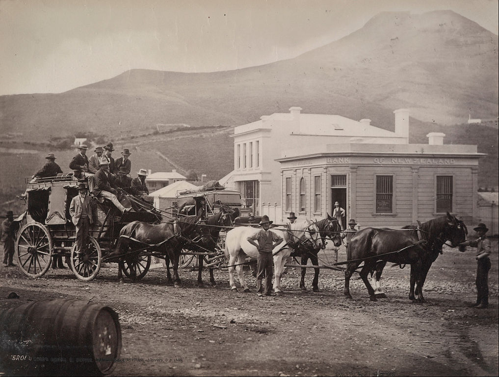 A coach full of people outside a white building. The coach is drawn by a team of horses. 